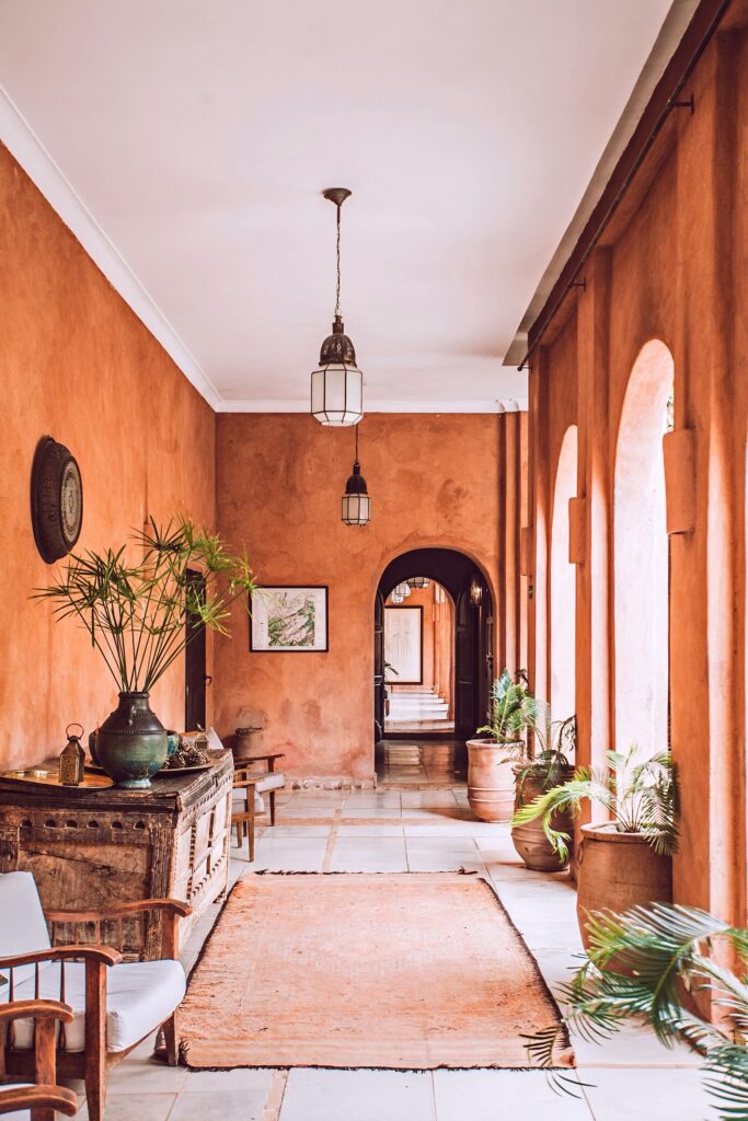 Está imagen diseñada para marketing. Es una pasillo de una casa que lleva a un patio interior, el pasillo tiene arcos redondos que dan al patio interior y deja entrar mucha luz, es de estilo marroquí con paredes de colores tierra, el pasillo está decorado con plantas y sillas.
