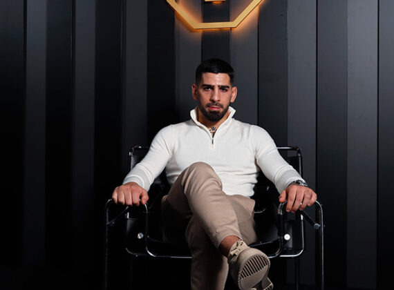 lia Topuria sentado en un sillón negro con ropa elegante de estilo de vida bajo el logotipo dorado de su marca personal.