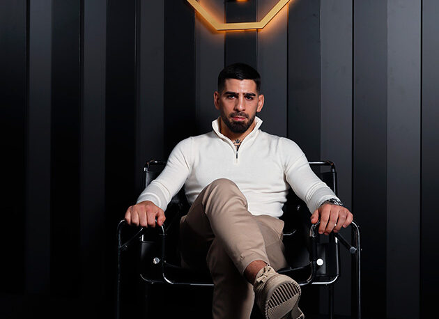lia Topuria sentado en un sillón negro con ropa elegante de estilo de vida bajo el logotipo dorado de su marca personal.