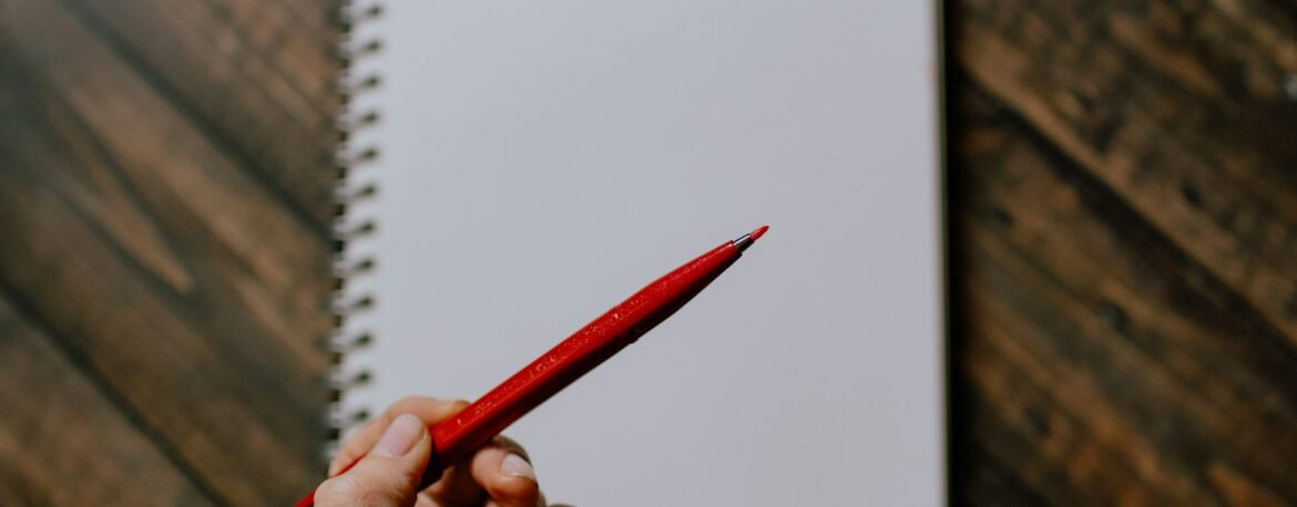 Una mano sostiene un bolígrafo rojo sobre un cuaderno en blanco encima de una mesa de madera